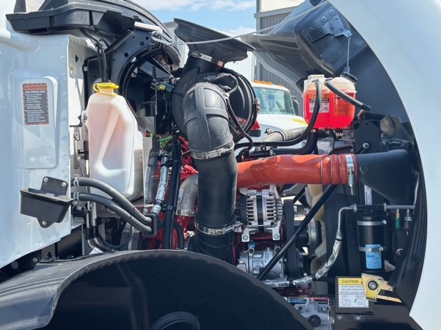 Front-left engine bay of a 2026 Mack MD6 truck showing coolant reservoir, large black intake hose, turbo/intercooler piping, alternator, and various hoses.
