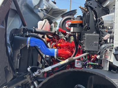 Engine bay of a Mack MD6 truck showing red engine block, blue coolant hose, various hoses, reservoirs, and electrical components.