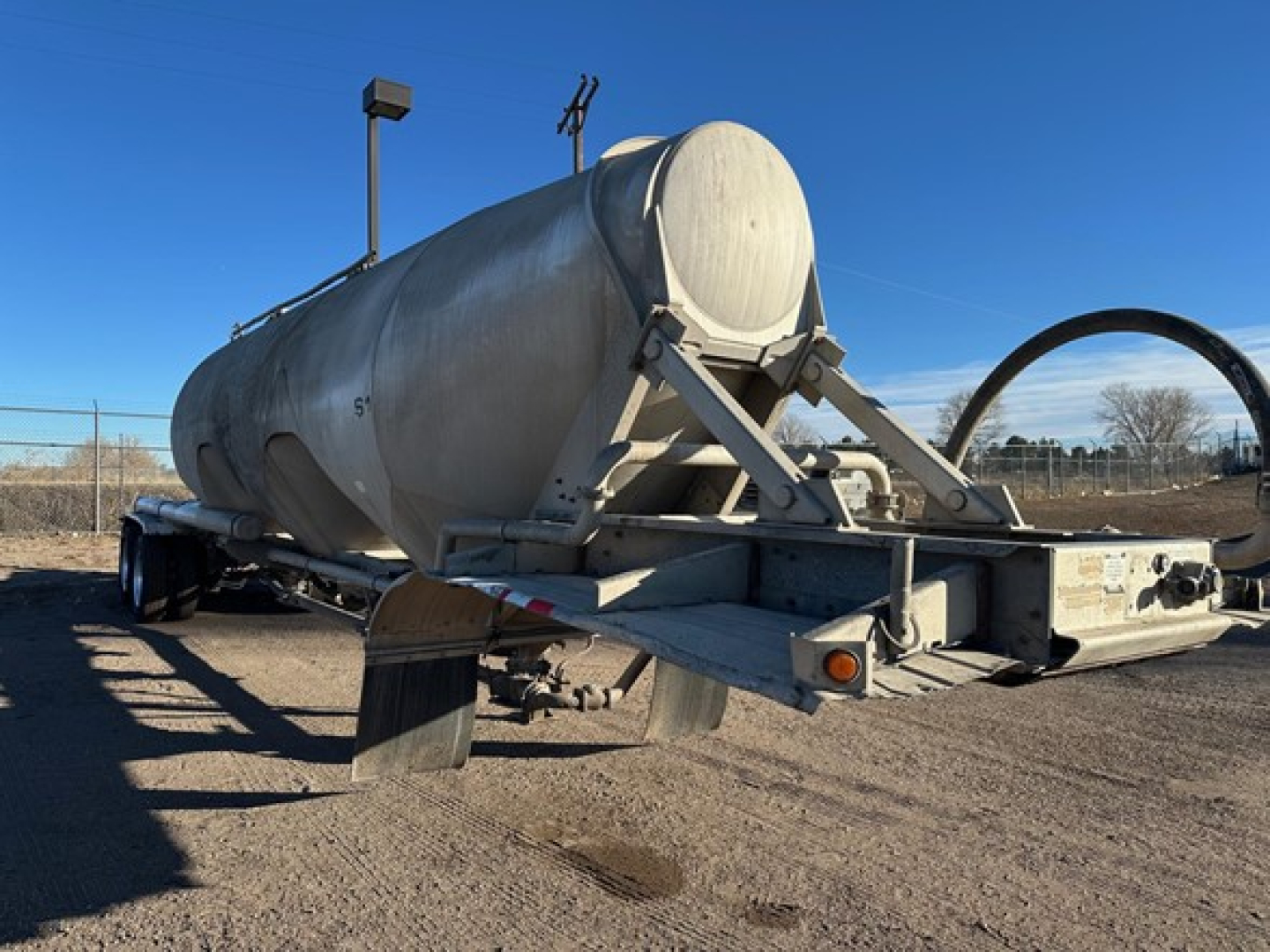 Chemical tanker trailer showing cylindrical tank on triple-axle chassis with rear loading chute and hoses.