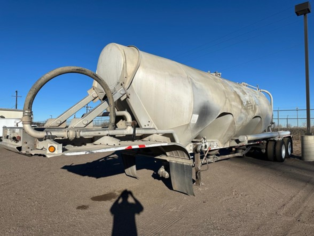 Pneumatic dry bulk trailer in service with cylindrical tank, rear chute and hoses visible. Side view shows tandem axles and support braces.