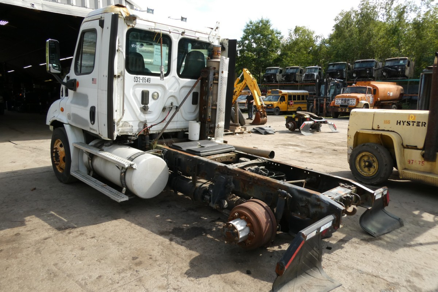 Truck chassis and cab of a Freightliner Cascadia daycab with detached front axle and exhaust, view from left side with industrial yard in background.