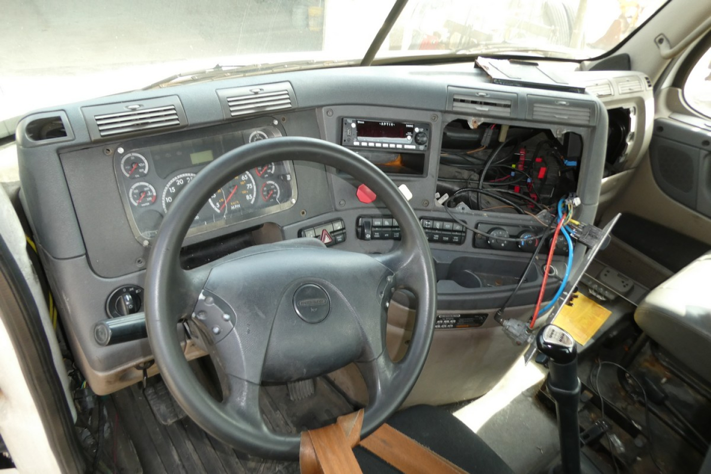 Cab interior of a Freightliner Cascadia 113 truck showing steering wheel, dashboard gauges, open instrument panel, exposed wiring, and missing center console panel.