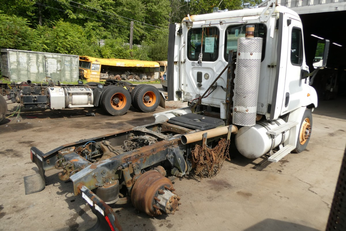 Truck cab of a white Freightliner Cascadia 113 with exposed drivetrain and fifth wheel, rusty rear axle components, in a yard.