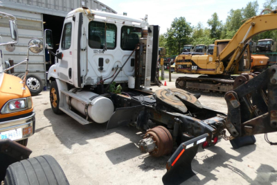 Freightliner Cascadia 113 tractor cab viewed from side with exposed drive shaft and mechanical components, mounted on a truck yard with construction equipment in background.