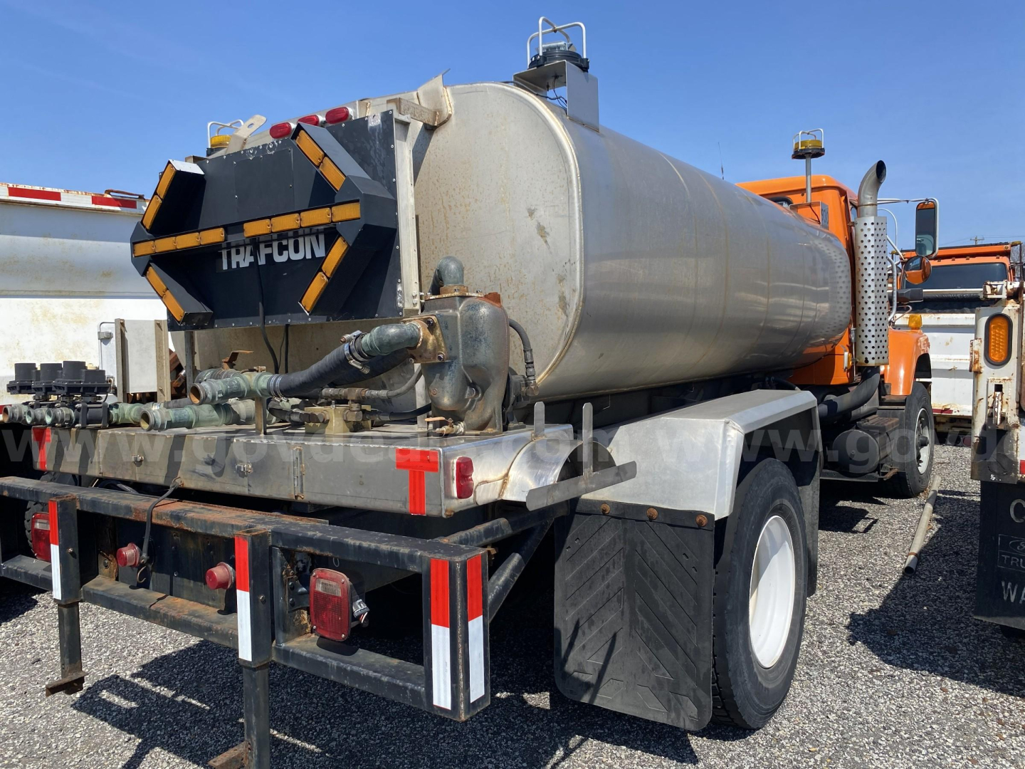 Tank trailer on a blue sky day, with large cylindrical stainless tank mounted on a truck chassis. Visible rear view shows hoses, valves, and a black-and-yellow hazard back panel labeled with ‘HICUN’ or similar branding.