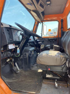 Truck cab interior showing steering wheel and driver's seat of an orange International truck. Dashboard with controls and gauges, pedals on floor, blue sky visible through window. Visible text includes faint watermark and dashboard labels.