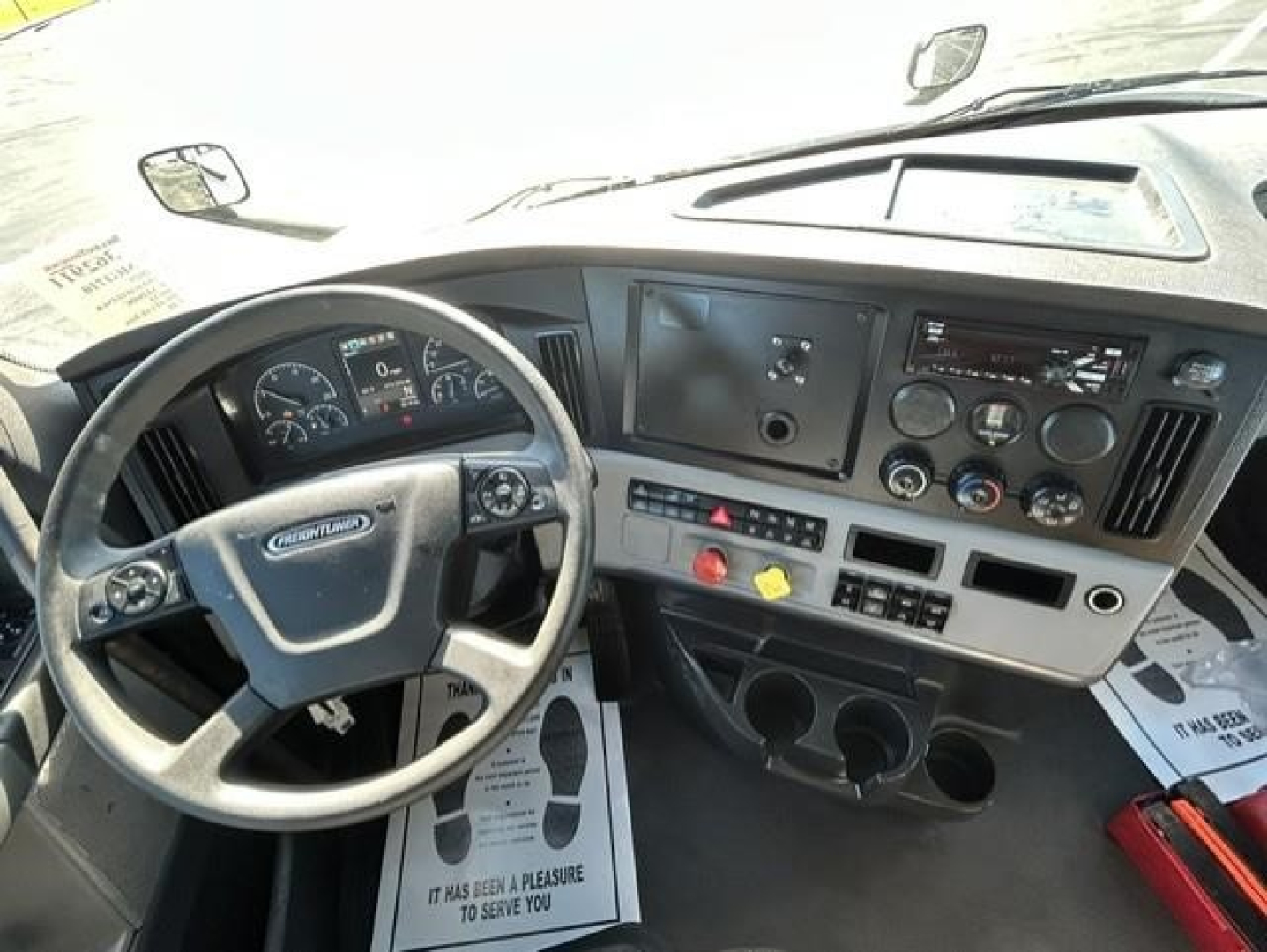 Dashboard and steering wheel of a Freightliner Cascadia cab showing steering wheel, instrument cluster, center console with gauges and controls, two cup holders, and AM/FM radio panel.