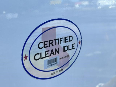 Certified Clean Idle sticker on a blue surface showing a circular seal with text CERTIFIED CLEAN IDLE and a small barcode, partially visible; background is part of a vehicle door.