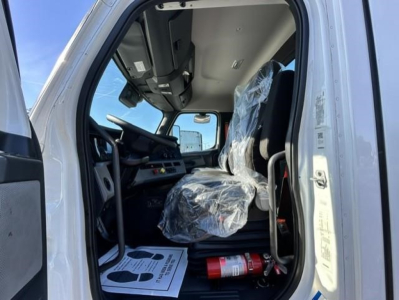 Truck cab interior of a used 2021 Freightliner Cascadia 126, driver's seat covered in plastic, steering wheel and dashboard visible, fire extinguisher on floor, sun light through open door