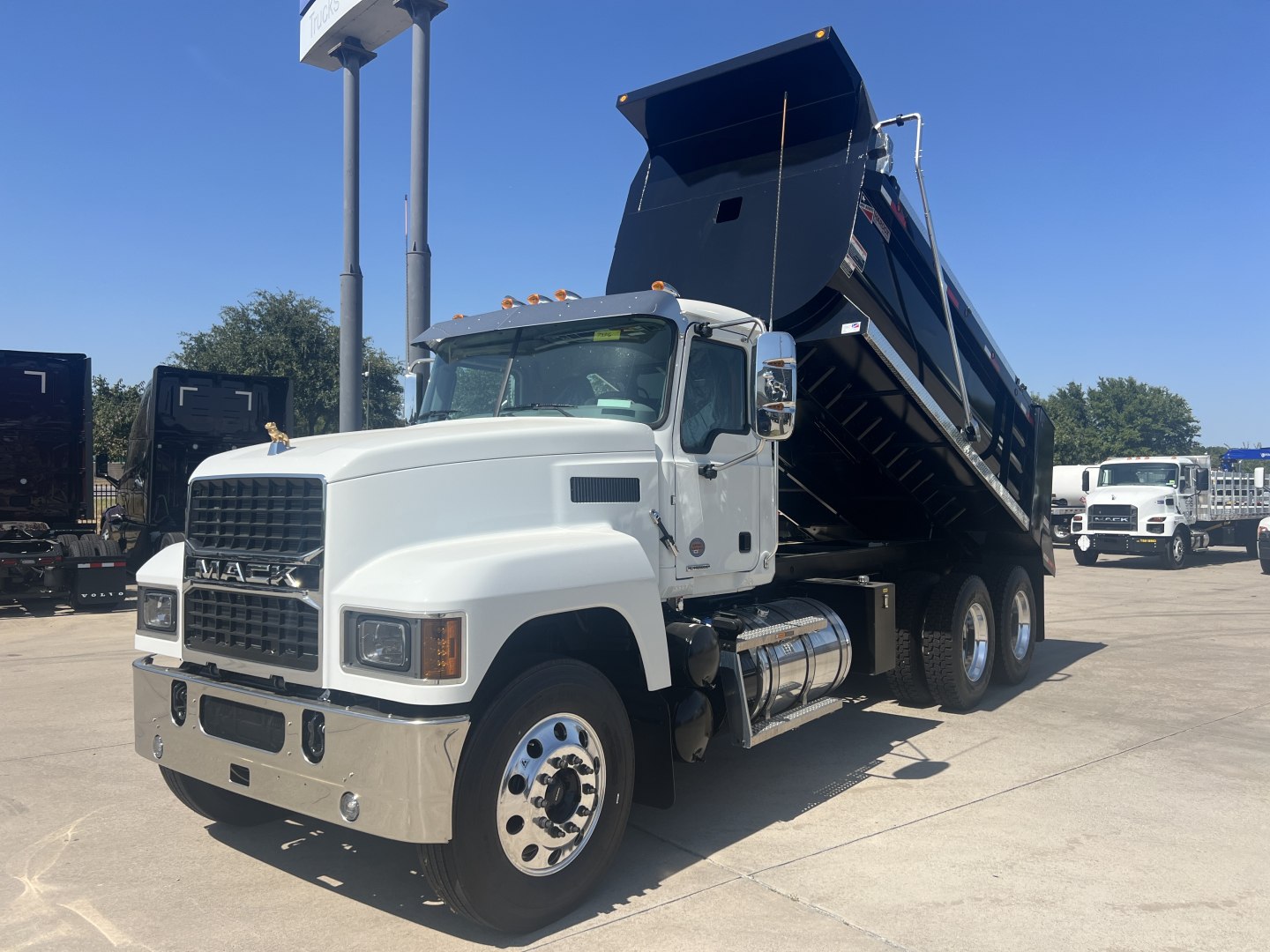 Dump truck in a dealership lot with a raised black dumping bed on a white Mack PI64 cab, front view showing chrome bumper and dual rear axles.