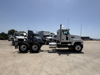Truck tractor, side view, white Mack PI64 with dual rear axles, fuel tank, and sleeper cab, parked on tarmac in dealership lot.