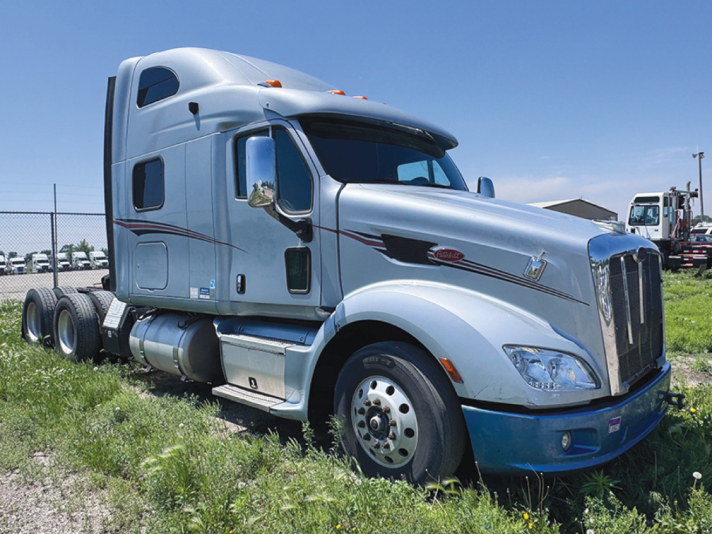 Peterbilt semi truck with silver cab in a grassy lot. Side shows fuel tanks and doors; no legible labels visible.