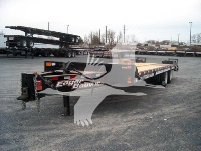 Flatbed trailer shown with the brand name Eager Beaver on the tongue, black frame, long deck, with stacked trailers in background.