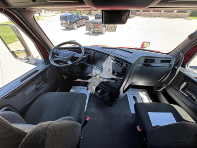 Truck cab interior of a Freightliner Cascadia 126 showing the dash, steering wheel, and front seats with a center console and paperwork on the passenger floor.