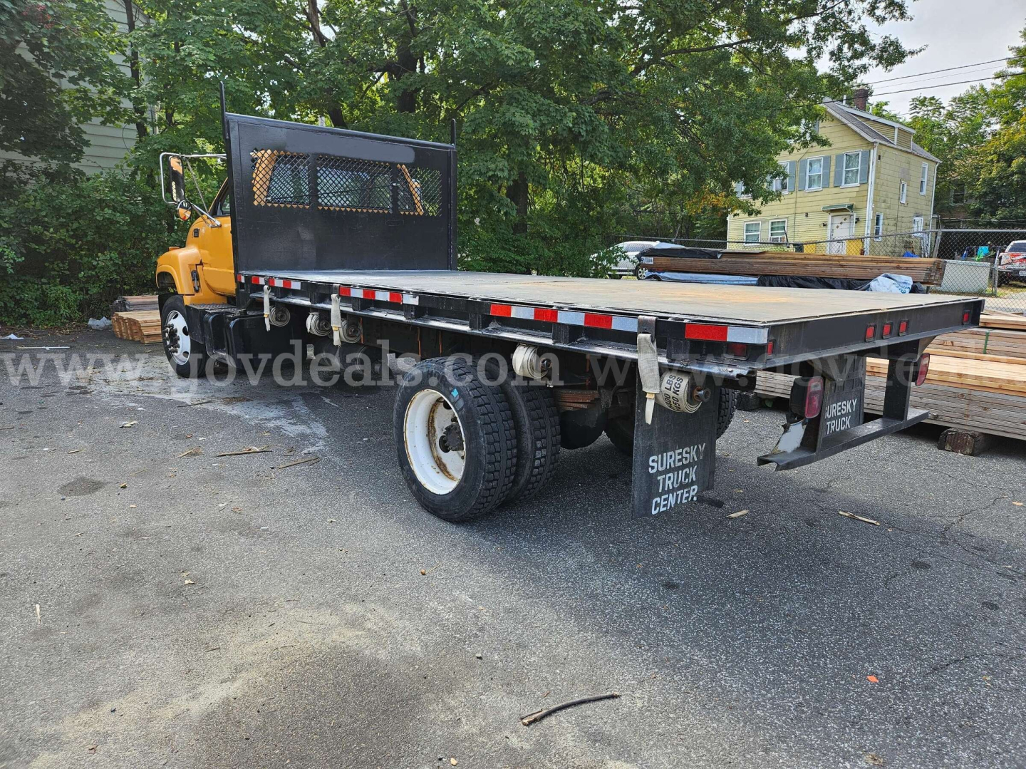 Flatbed tow truck with yellow cab and long aluminum flatbed, rear view showing dual rear wheels and black headache rack, parked on asphalt.