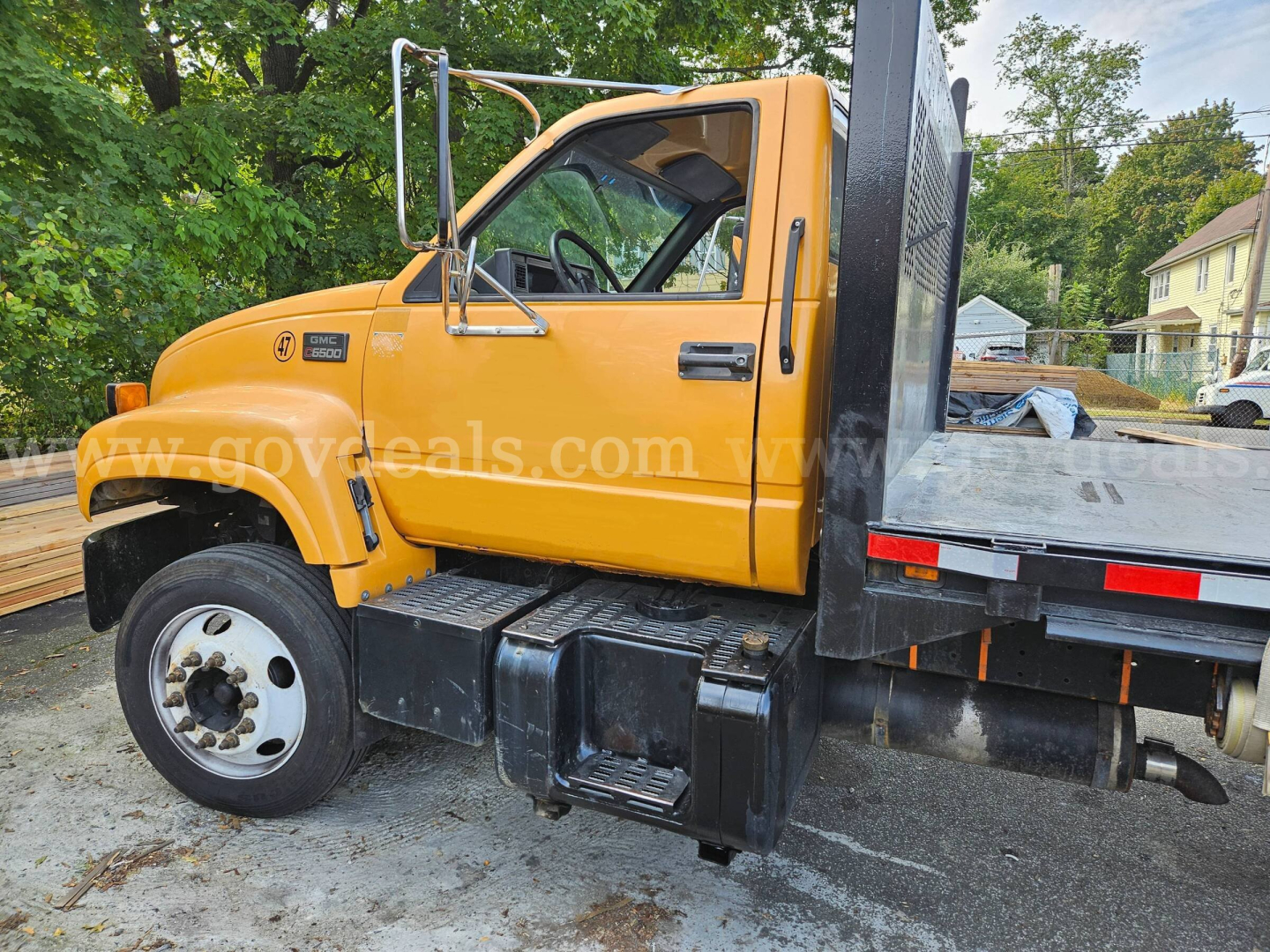 GMC C6500 flatbed dump truck in orange cab, side view showing wheel, door, fuel/air intake, and flatbed with metal steps.