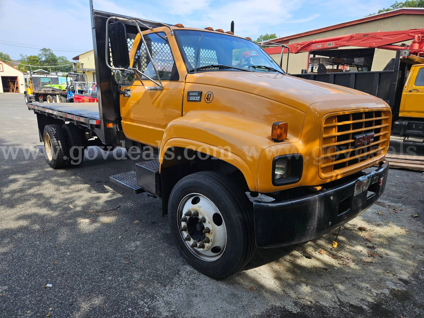 GMC C6500 flatbed tow truck in service orange/yellow with flatbed, front grille, and side steps; visible text includes GMC logo and model badge; truck shows VIN tag area not fully legible.