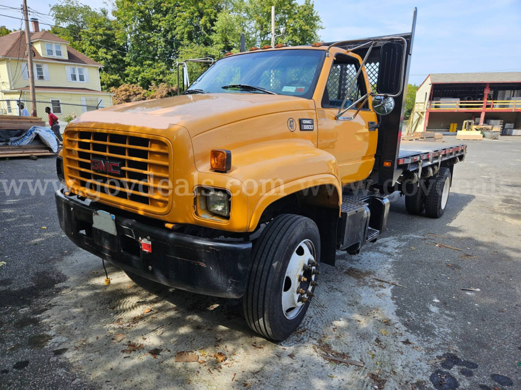 GMC C6500 flatbed dump truck in orange showing front grille and dual rear axles with cab doors open