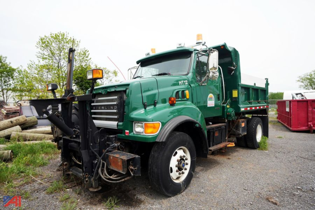Green Sterling L9500 dump truck with wing and plow attachment at front, cab with orange warning lights, and dump bed in background.