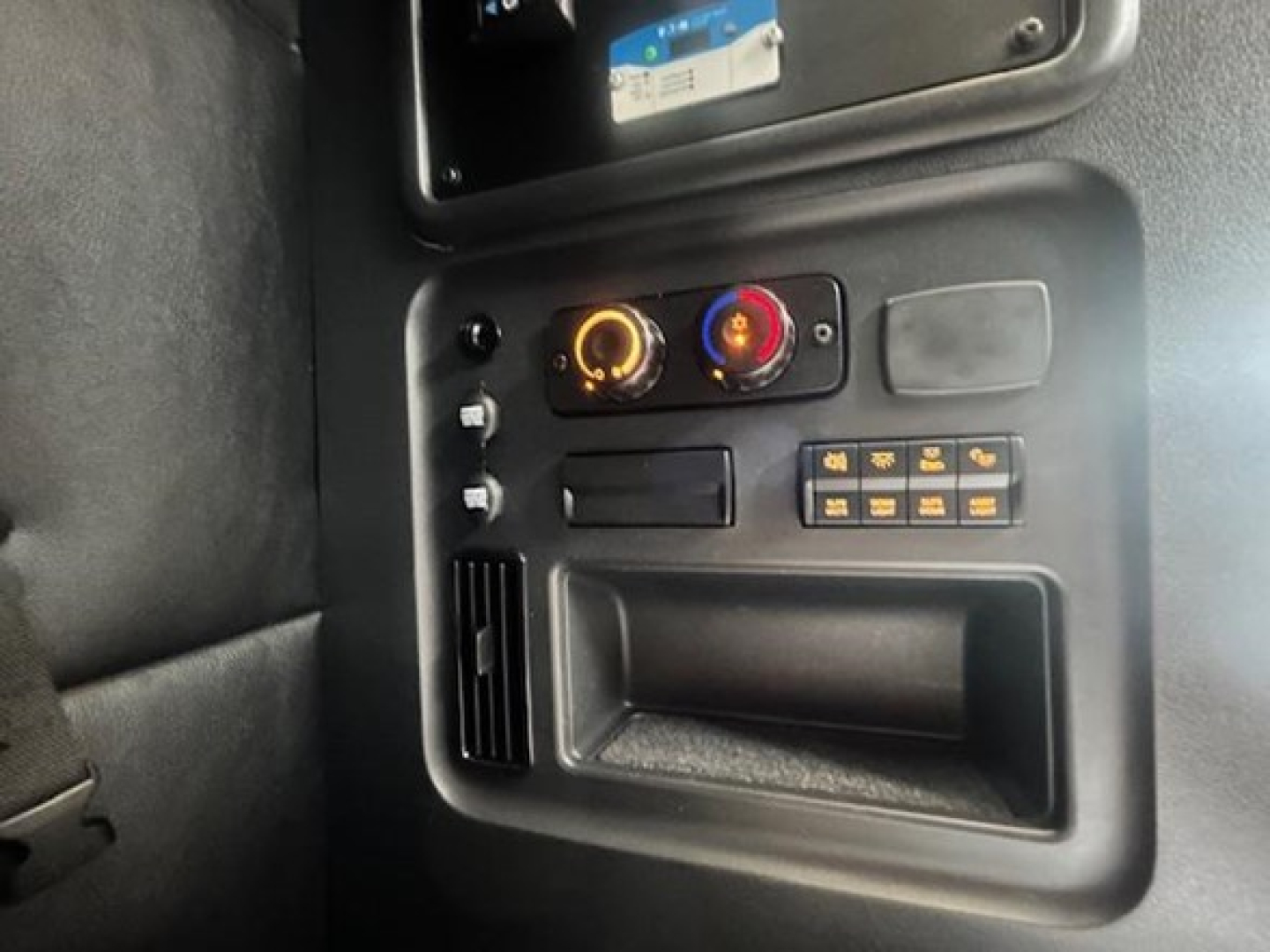 Vehicle cab interior showing control panel with circular dials, small display, and storage pocket in the center console.