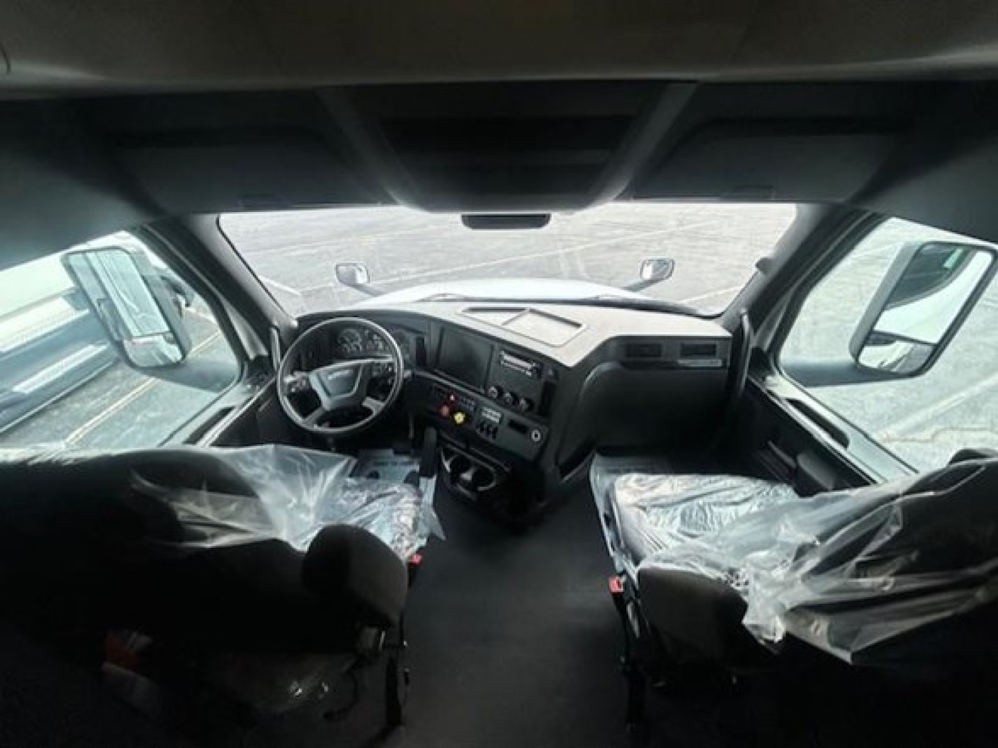 Truck cabin interior of a 2023 Freightliner Cascadia 126, front seats wrapped in plastic, dashboard with steering wheel and controls visible.