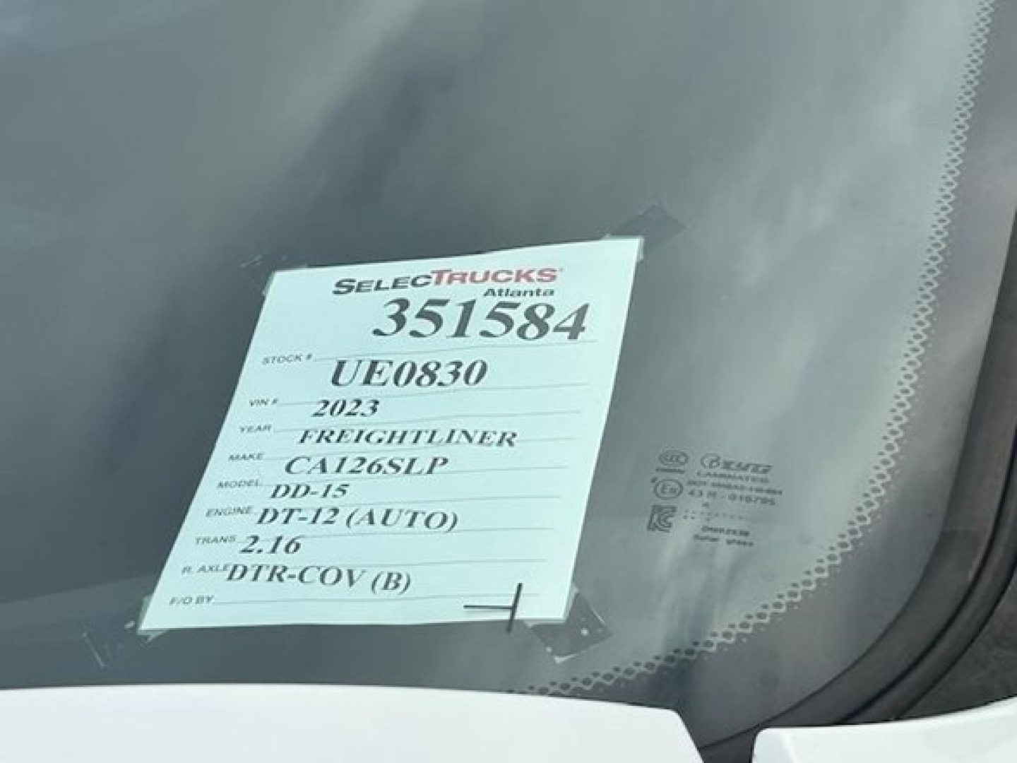 Truck windshield with a sticker showing: STOCK # UF0830, YEAR 2023, MAKE Freightliner, MODEL DD-15, MT. 2.16, ENGINE DT-12 (AUTO), DTR-COV (B), VIN 3AKJHHDR6PSUE0830, stock 351584.