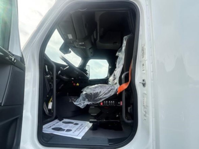 Truck cab interior of a 2023 Freightliner Cascadia 126, driver seat with plastic cover, steering wheel, dashboard and pedals visible from open door.