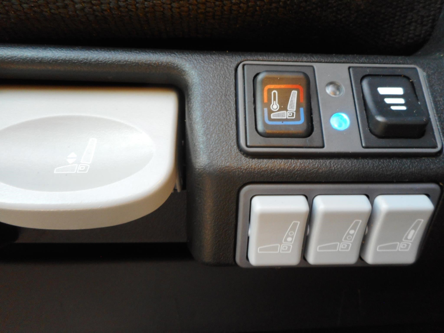 Storage compartment area showing seat heater switch with orange indicator and three gray control buttons with seat icons; a large beige circular cover knob on left; part of dashboard panels.