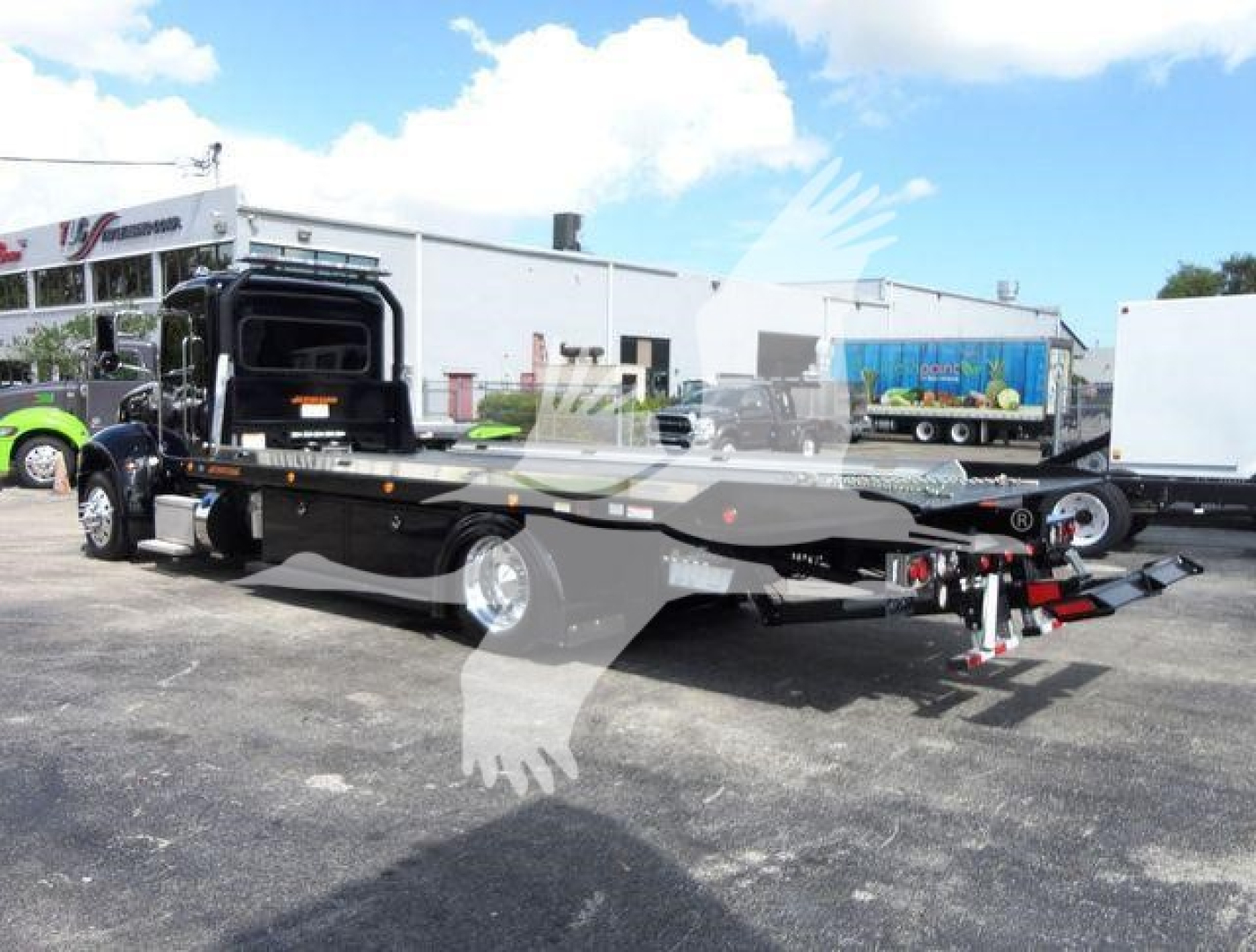 Peterbilt 337 rollback wrecker truck in a dealership lot, flatbed with ramps, black color, chrome wheels, background buildings and trailers.