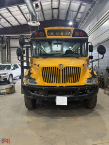 Front view of a yellow 2020 IC Corporation CE Series school bus inside a workshop, showing grille, bumper, headlights, and 'SCHOOL BUS' sign above windshield.