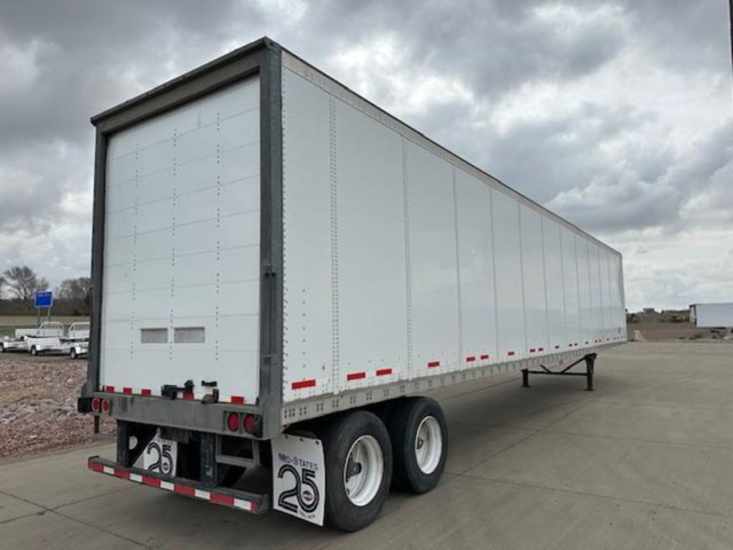 Back/side view of a white semi-trailer with tandem axles, showing rear doors and taillights; small red reflectors along lower side; license-plate-like numbers on mud flaps reading 25.