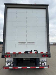 Rear view of a white Wabash trailer with closed doors, black frame, red and white reflective tape, and taillights.