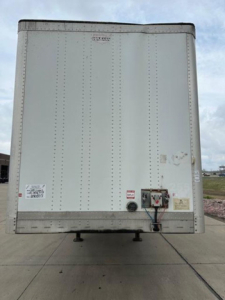 Rear view of a white Wabash trailer with doors closed showing various safety labels and a connector; small black and red manual crank near bottom right; no visible branding text legible.