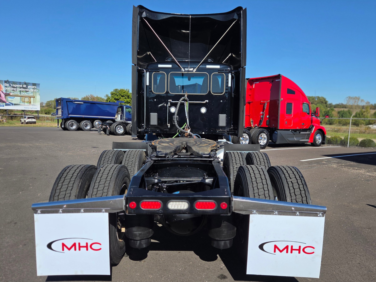 Peterbilt 579 tractor rear view showing fifth wheel, dual rear axles, and mud flaps with MHC logo. Cab raised with sleeper, other trucks in lot.