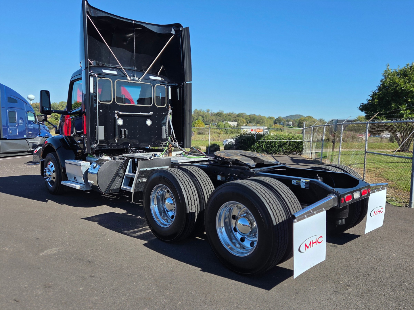 Peterbilt 579 tractor with raised hood, dual rear axles, chrome fuel tanks, and white MHC mud flaps in a dealership lot.