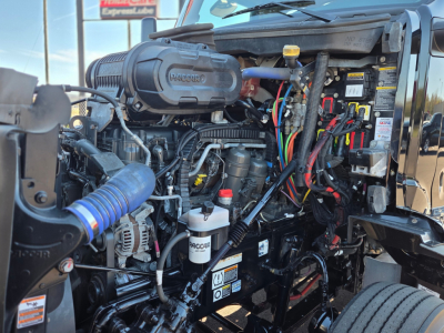 Peterbilt 579 engine bay showing Cummins or PACCAR engine with various hoses, wiring harnesses, and fluid reservoirs visible.