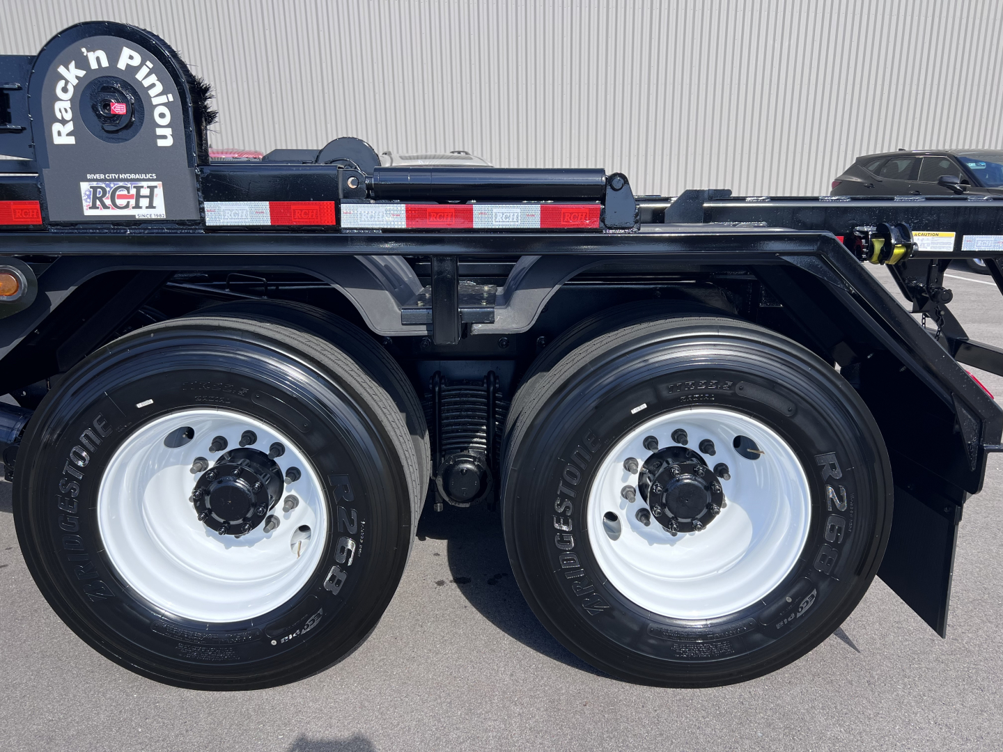Truck wheel area showing dual rear axles with Bridgestone tires and white steel rims. Left rear tire shows 'BRIDGESTONE' branding and the wheel well shows suspension components. A spare wheel mount with 'Rack'n Pinion' label partially visib