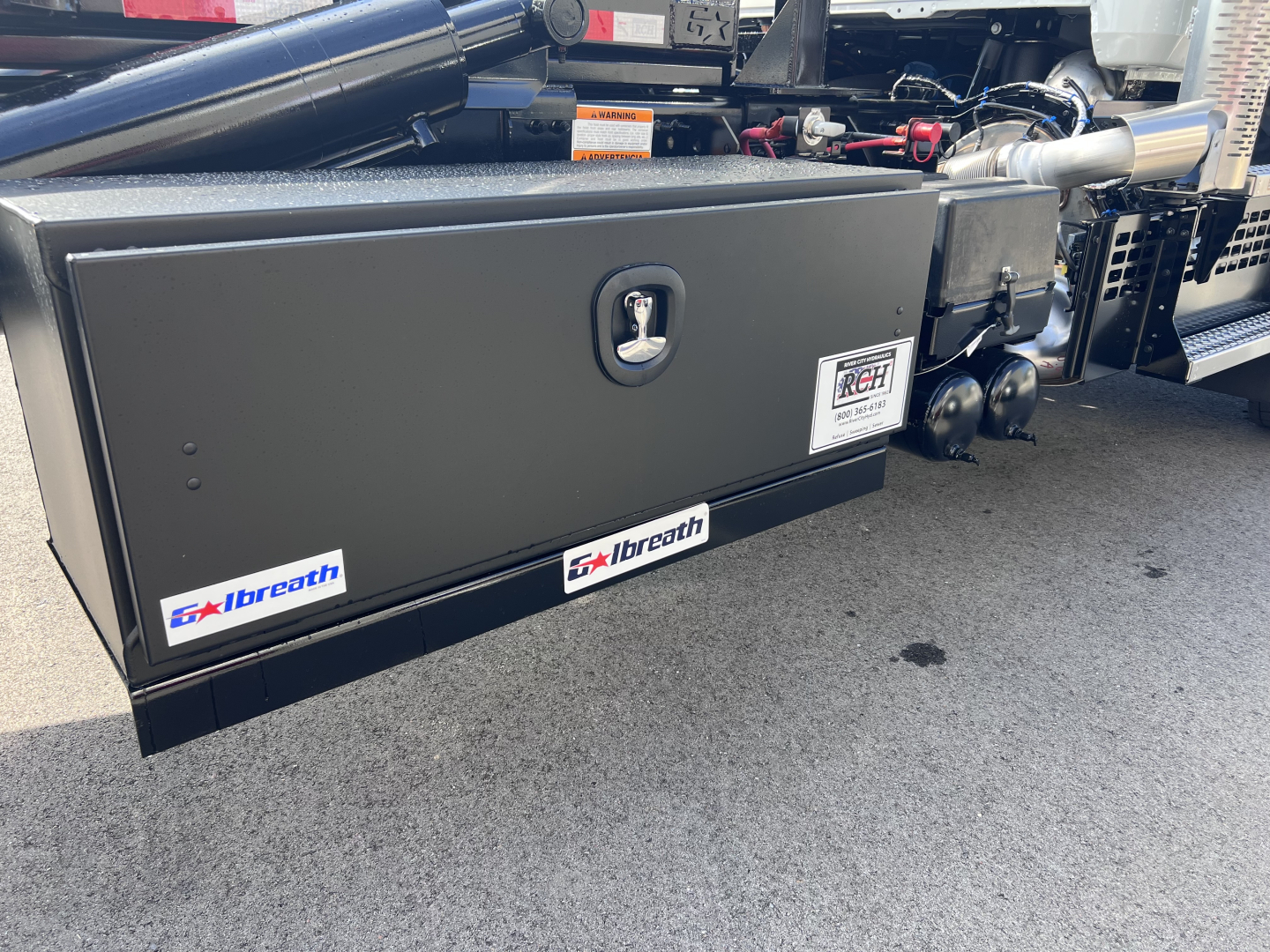Truck component showing storage box on Mack GR64B roll-off with toolbox lid and badges reading 'GxLbreath' and mechanical piping nearby