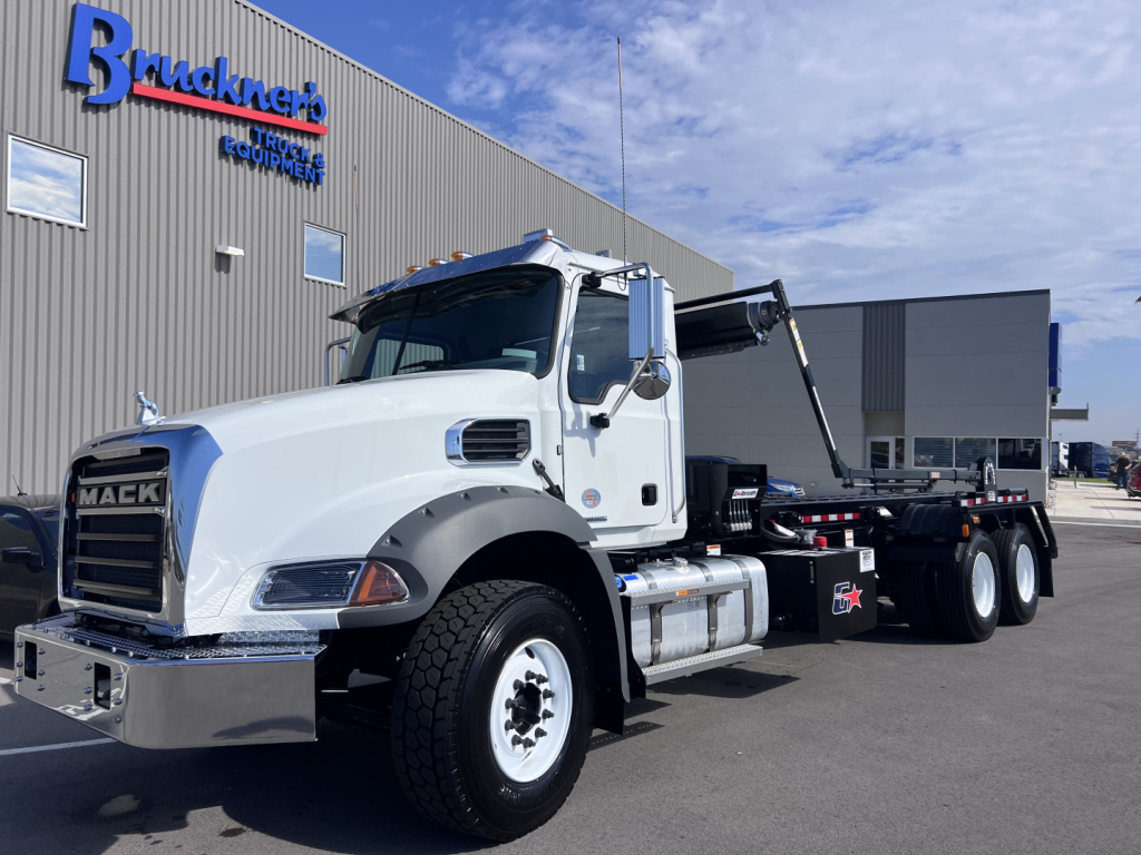 New 2026 Mack GR64B roll-off truck, white cab, in-service with Brukner's Equipment backdrop. Visible text: Mack grille, GR64B model, 6x4 undercarriage, chrome bumper, fuel tanks, tool compartments, and hydraulic boom. VIN 1M2GR4GC3TM053859,