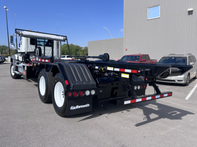 Mack GR64B roll-off truck showing left rear view with dual rear axles, black chassis, white wheels, taillights, and reflective red/white safety stripes.