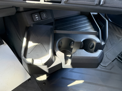 Cab interior center console shown with dual cup holders and storage tray in a new Mack PR64T76H truck. Includes small compartments and a raised step; plastic bin with circular cup slots.