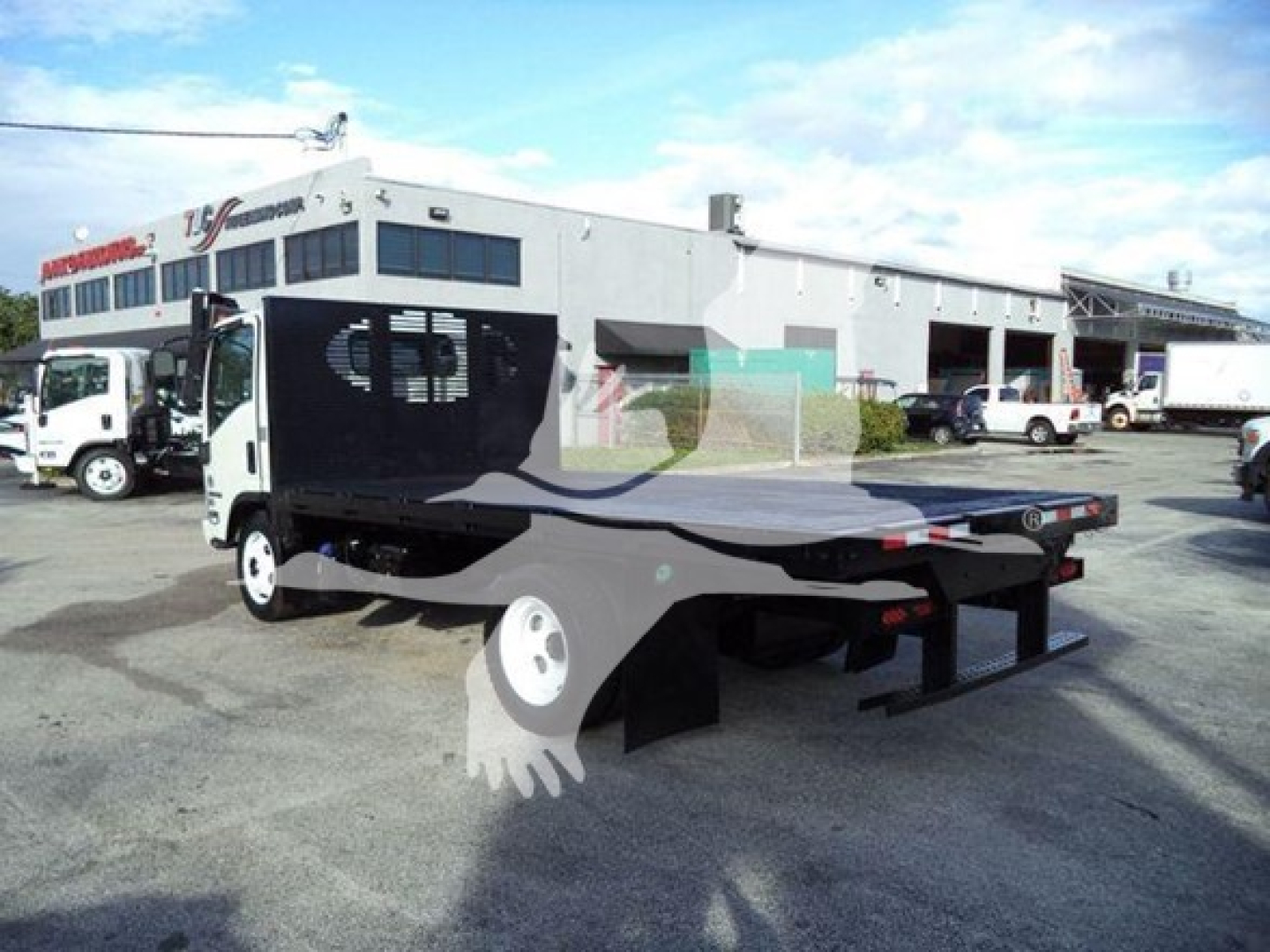 Isuzu NRR flatbed truck in used condition with a drop bed and hydraulic tilt mechanism, side cab visible, dealership in background.