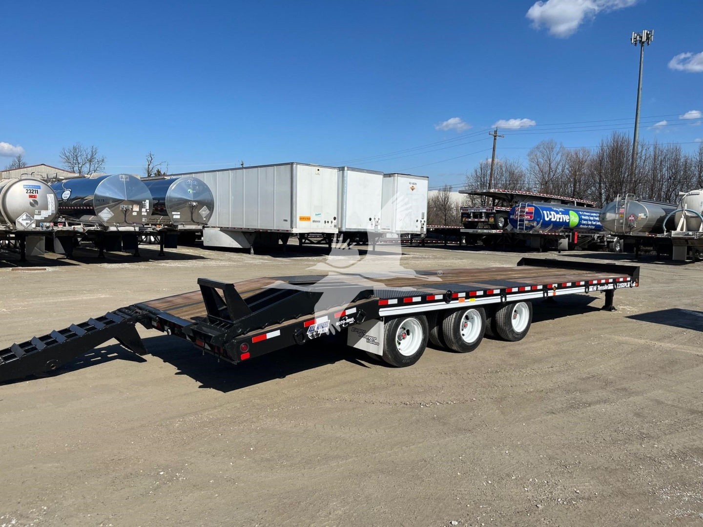 Trailer showing a low-profile gooseneck flatbed trailer with triple axles and loading ramps, in a dealership lot.