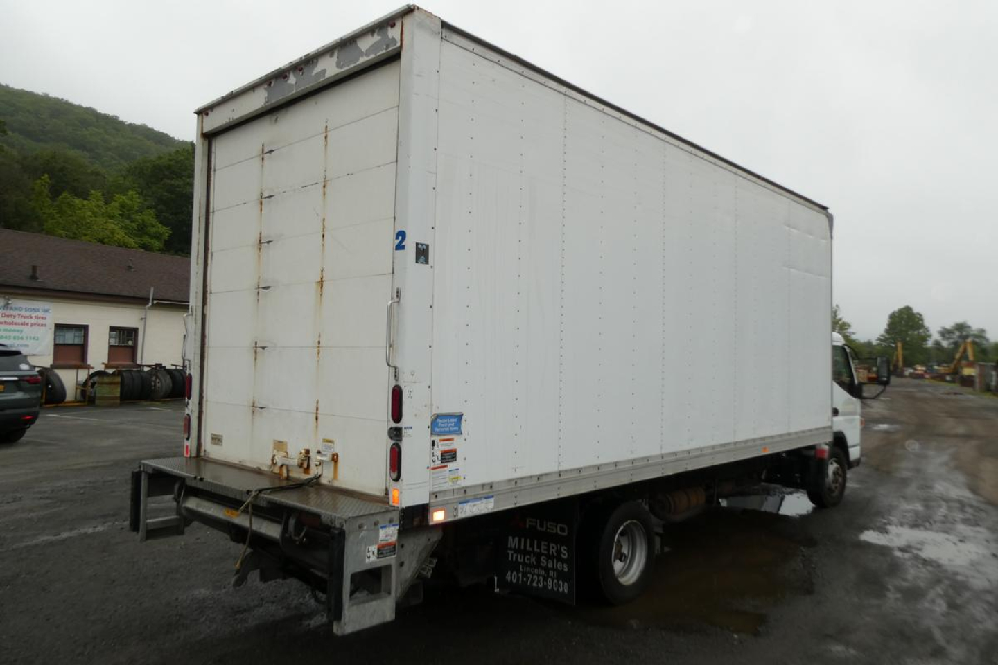 Truck box rear and side view showing white cargo box on Mitsubishi Fuso Canter FE140. Visible decals near rear include small stickers and taillights; chassis and wheels underneath.