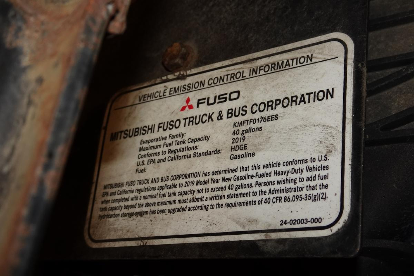 Vehicle emission control information plate on Mitsubishi Fuso Canter; label shows Mitsubishi Fuso Truck & Bus Corporation, model details, fuel type gasoline, and regulatory text.