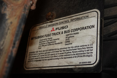 Vehicle emission control information plate on Mitsubishi Fuso Canter; label shows Mitsubishi Fuso Truck & Bus Corporation, model details, fuel type gasoline, and regulatory text.