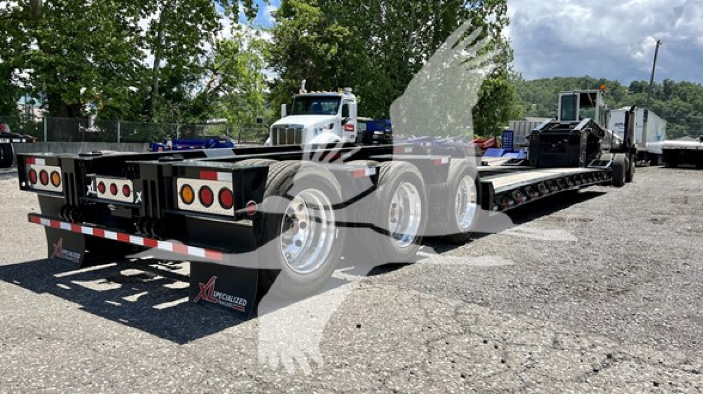 Lowboy trailer showing multiple axles and rear lights; XLM Coast-to-Coast logo overlay; metal ramps and mud flaps visible.