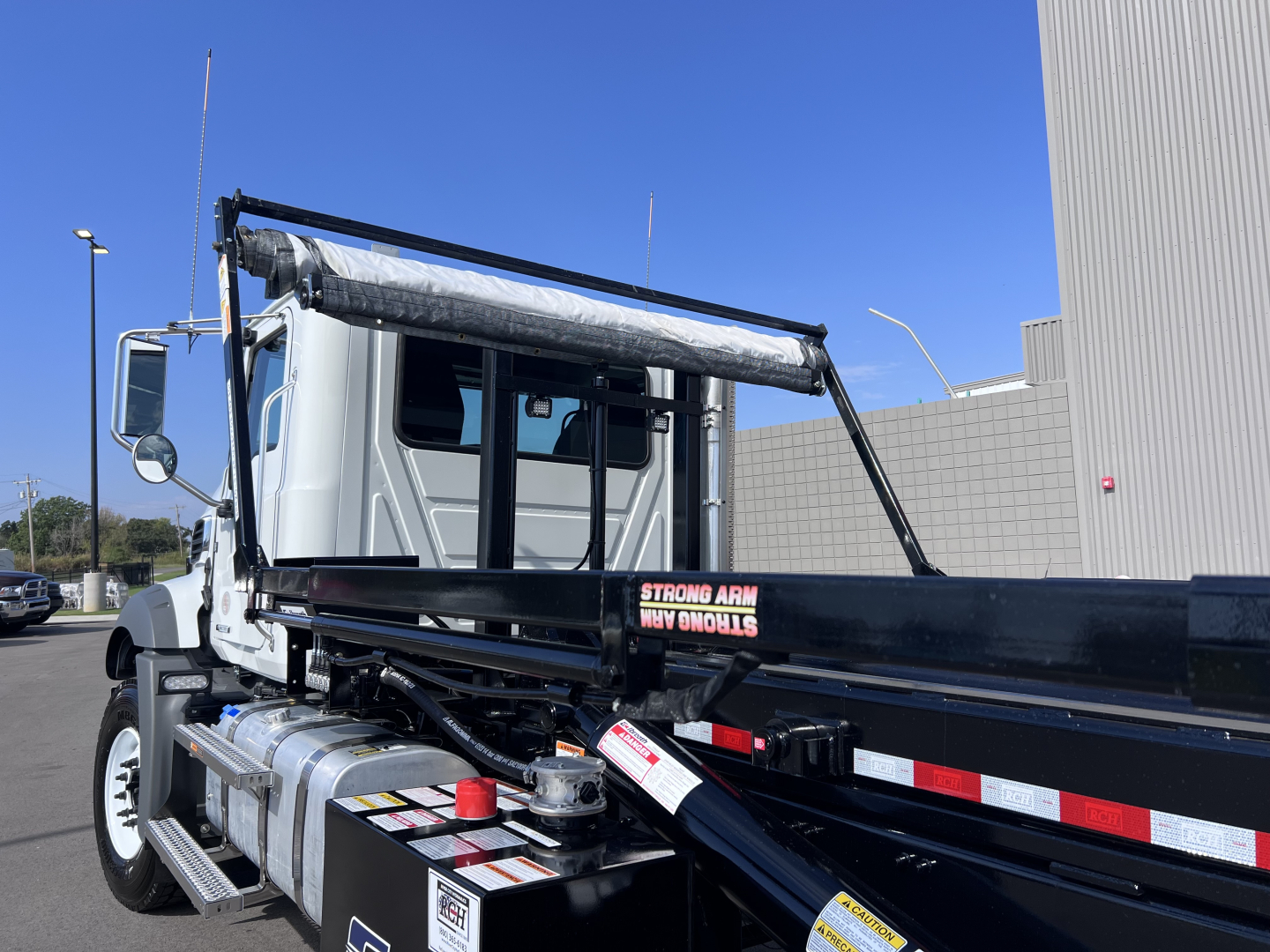 New Mack GR64B roll-off truck showing cab, long black arm with white and red reflectors, and metal fuel tank. Visible VIN 1M2GR4GCXTM053857 and stock 70288.
