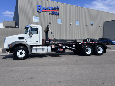 Mack GR64B roll-off truck shown in side view with white cab and long black chassis in front of a dealership building.