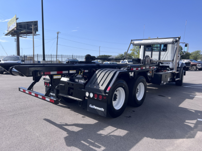 Mack GR64B rolloff truck rear view showing dual rear axles, aluminum flatbed frame, black mud flaps, and rear lighting; side text not visible.